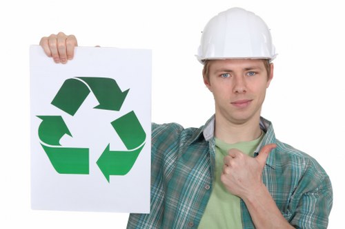 A young man wearing a white safety helmet and a casual checked shirt, standing outdoors on what appears to be a paved or concrete surface. He is holding a white sign featuring a large green recycling symbol composed of three arrows forming a triangle, which is prominently displayed in front of him. The man's facial expression is neutral, with a slight, confident smile, and he is giving a thumbs-up gesture with his right hand. The background is plain and uncluttered, focusing on the man and the sign. This image visually emphasizes environmental responsibility and waste recycling themes relevant to commercial rubbish and waste removal services in Catford, suggesting a professional approach to sustainable waste management as offered by Commercial Waste Catford.