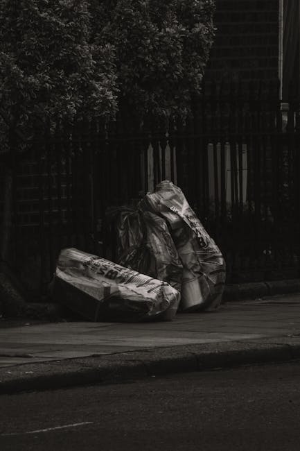 Three large black plastic garbage bags, filled and tied at the top, are placed on the pavement next to a black metal fence, with a row of dense, leafy trees behind them. The bags appear to contain hou