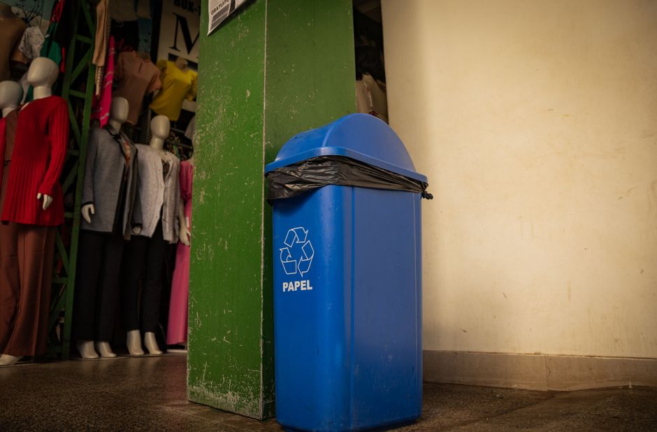 A row of five brightly coloured wheelie bins aligned against a plain, light blue wall with a brick base. The first bin on the left is yellow with a white label reading 'RACCOLTA CARTA,' containing som