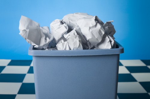 A light blue plastic rubbish bin filled with crumpled white paper and shredded paper, placed on a black and white checkered tiled floor. The background features a solid blue wall, suggesting an indoor or outdoor setting typical of waste collection areas in Catford. The crumpled papers in the bin display various textures with visible creases and folds, indicative of used packaging or discarded documents. The bin is positioned centrally in the image, with the paper content overflowing slightly at the top, emphasizing its fullness. The environment appears clean and orderly, consistent with commercial rubbish collection practices by companies like Commercial Waste Catford. This visual highlights the importance of proper waste management and disposal of paper and packaging waste, which is part of the waste removal services offered within the London borough of Lewisham, near Catford postcodes.